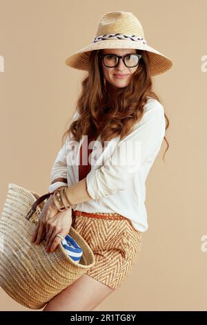 Strandurlaub. Eine lächelnde, trendige Frau in weißer Bluse und Shorts vor beigefarbenem Hintergrund in Brille mit Strohtasche und Sommerhut. Stockfoto