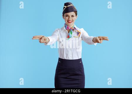 Lächelnde, stilvolle Stewardess vor blauem Hintergrund in Uniform, die zeigt, wo die Ausgänge sind. Stockfoto