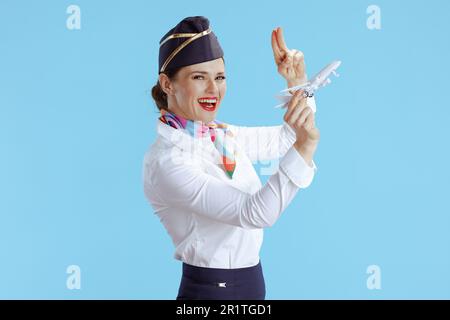 Glückliche, stilvolle Flugbegleiterin auf blauem Hintergrund in Uniform mit einem kleinen Flugzeug salutiert. Stockfoto