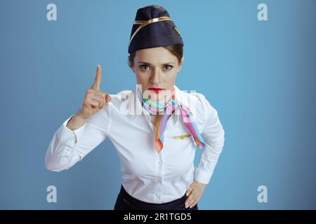 Moderne weibliche Stewardess mit blauem Hintergrund in Uniform, mit erhabenen Fingern, die Aufmerksamkeit erregen. Stockfoto