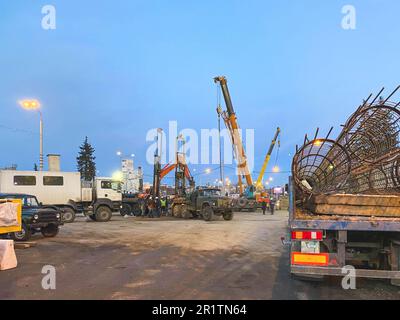 Bau einer gebrochenen Brücke an einer verkehrsreichen Straße. Die Baumaschinen auf dem Gelände errichten Betonblöcke. Der Lkw trägt einen Metallrahmen. Stockfoto