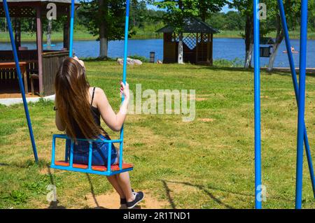 Eine wunderschöne junge Frau mit langen Haaren fährt im Sommer auf einer Schaukel im Park auf der Natur am Ufer des Sees. Stockfoto