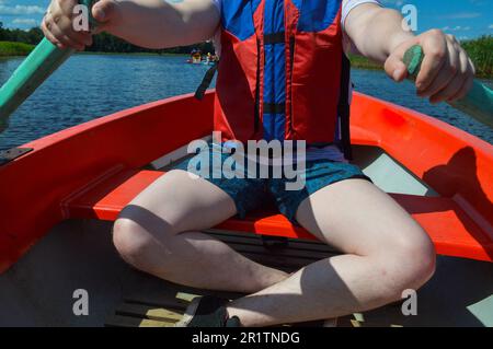 Ein Mann in einem roten Boot, Shorts und einer Schwimmweste paddelt mit Rudern auf einem Boot für einen Spaziergang auf dem Wasser des Flussufers in der Natur. Stockfoto