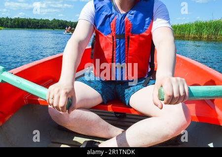 Ein Mann in einem roten Boot, Shorts und einer Schwimmweste paddelt mit Rudern auf einem Boot für einen Spaziergang auf dem Wasser des Flussufers in der Natur. Stockfoto