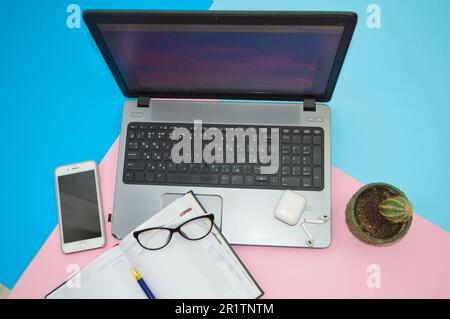 Arbeitsplatz zu Hause, Heimbüro für Fernarbeit mit einem Computer, Laptop, Mobiltelefon, Smartphone, Notizblock, Kopfhörer und Brille auf einem pinkfarbenen und blauen Ba Stockfoto