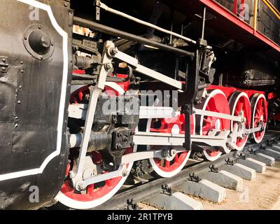 Große Eisenräder eines rot-schwarzen Zuges, der auf Schienen und Aufhängungselementen steht, mit Federn einer alten industriellen Dampflokomotive. Stockfoto