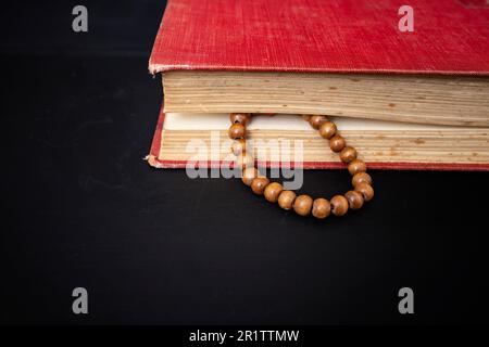 Holzperlen an einer Schnur zwischen den Seiten eines roten, harten Eindeckbuchs, Nahaufnahme auf dunklem Hintergrund. Gebetskonzept Stockfoto