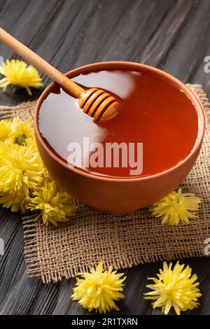 Honig in einer Tonschüssel, Dipper und gelbe Blumen auf Textil- und dunklem Holzhintergrund. Ein Honigstock liegt in einer Keramikschüssel mit Honig Stockfoto