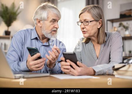 Fokus auf schwarze Smartphones, die ältere Männer und Frauen in Business-Freizeitkleidung im Innenbereich des Home-Office halten. Professionelle Partner tauschen Ideen über neue Strategien am entfernten Arbeitsplatz aus. Stockfoto