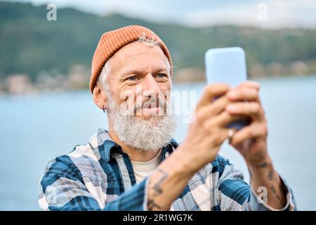 Glücklicher älterer Hipster, der im Naturpark steht und Fotos auf dem Smartphone macht. Stockfoto