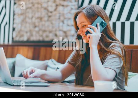 Eine ziemlich lächelnde, beschäftigte junge Frau, die im Café sitzt und moderne Geräte kommuniziert Stockfoto