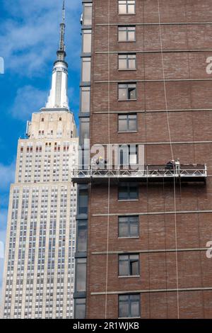 Die Arbeiter führen auf einer abgehängten Plattform Ziegelumschläge durch, wobei das Empire State Building im Hintergrund, 2023, New York City, USA, steht Stockfoto