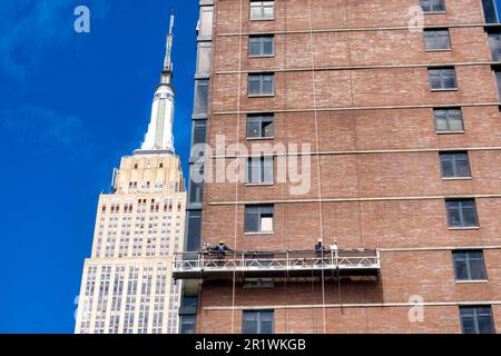 Die Arbeiter führen auf einer abgehängten Plattform Ziegelumschläge durch, wobei das Empire State Building im Hintergrund, 2023, New York City, USA, steht Stockfoto