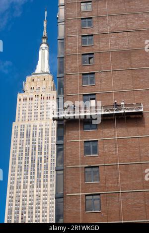 Die Arbeiter führen auf einer abgehängten Plattform Ziegelumschläge durch, wobei das Empire State Building im Hintergrund, 2023, New York City, USA, steht Stockfoto