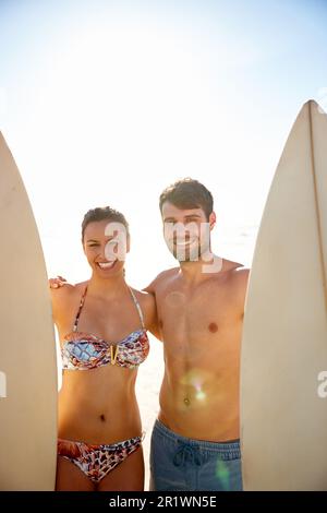 Gehen wir da raus. Porträtaufnahme eines glücklichen jungen Paares, das mit seinen Surfbrettern am Strand lächelt. Stockfoto