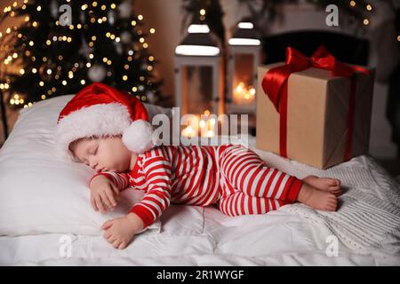 Baby im Weihnachtsschlafanzug und Weihnachtsmannmütze schlafen in der Nähe der Geschenkbox auf dem Bett drinnen Stockfoto