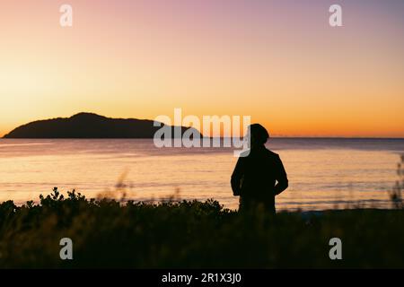 Silhouette eines Mannes, der bei Sonnenaufgang auf einer Sanddüne mit Blick auf Jimmy's Beach steht. Hawks Nest im Zentrum von NSW Australien Stockfoto