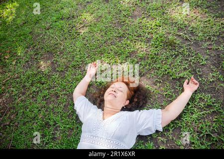 Reife hispanische Frau, die auf dem Rasen liegt, mit geschlossenen Augen lächelt, die Arme entspannt hält und einen Moment der Ruhe genießt. Stockfoto