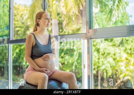 Schwangere Frau, die zu Hause auf Sportball trainiert. Schwangere Frau, die Entspannungsübungen mit einem Fitness-Pilates-Ball macht. Vor dem Hintergrund des Fensters Stockfoto