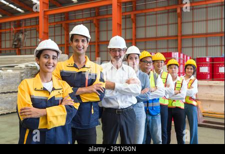 Eine Gruppe von männlichen und weiblichen Fabrikarbeitern lächelt zusammen mit den Armen in der Fabrik. Alle tragen Schutzkleidung und Helm. Ich Habe Angst Stockfoto