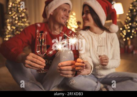 Ein Paar in Weihnachtsmannhüten mit Glitzer und Champagnergläsern, konzentriert euch auf das Feuerwerk. Weihnachtsfeier Stockfoto