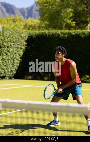 Ein birassistischer Mann mit Schläger, der darauf wartet, dass der Ball auf einem sonnigen Tennisplatz im Freien zurückkehrt, Kopierraum. Sommer, gesunder Lebensstil, Sport, Hobbys und Urlaub Stockfoto