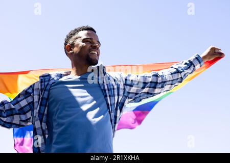 Fröhlicher afroamerikanischer junger schwuler Mann mit Regenbogenfahne, die Arme gegen den klaren Himmel streckt Stockfoto