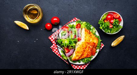 Gebratenes weißes Fischfilet mit Gemüsesalat aus Salat, Kirschtomaten und roten Zwiebeln, schwarzer Tischhintergrund, Draufsicht Stockfoto