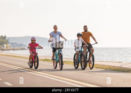 Zwei Kinder mit Helmen auf dem Kopf und lächelnde Eltern, die auf einer familienfreundlichen Radroute entlang der Küste mit Blick auf die Küste Fahrräder fahren. Stockfoto