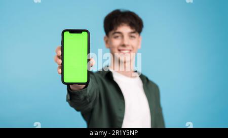 Ein junger Mann lächelt und zeigt sein Handy mit grünem Bildschirm auf blauem Hintergrund in die Kamera Stockfoto