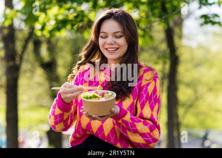 Blick auf eine lächelnde Frau, die eine Schüssel mit Gemüse, Obst, Garnelen und Reis mit Essstäbchen isst Stockfoto