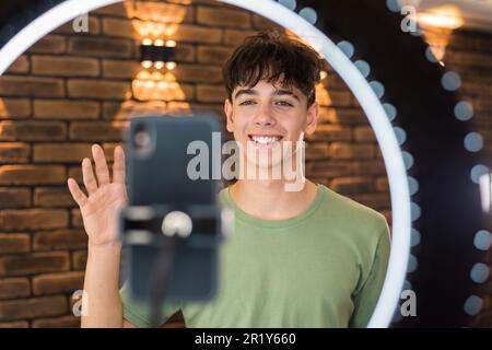 Ein junger lächelnder Mann Blogger Influencer, der sich selbst auf einem Smartphone auf einem Stativ mit Ringlicht in einem Studio fotografiert hat Stockfoto