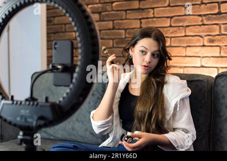 Eine junge Bloggerin, die sich schminkt und sich mit einem Smartphone auf einem Stativ mit Ringlicht in einem Studio fotografiert Stockfoto