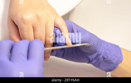 Maniküre Formen, feilen die Nägel einer schönen Frau, Finger halten. Ein weißer Manikürmeister arbeitet in Handschuhen an einem weißen Arbeitsplatz im Wellnesssalon. Weich Stockfoto