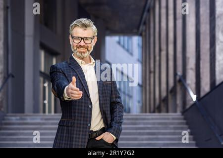 Reifer, erfolgreicher, grauhaariger Geschäftsmann, der lächelt und in die Kamera schaut, die Hand in der Hand hält, Grußgeste, erfahrener Boss. Stockfoto