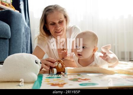 Eine fürsorgliche, fröhliche Mutter spielt mit ihrem kleinen Kind und liegt auf dem Boden in einem hellen Kinderzimmer. Verschiedene Spielzeuge, ein Buch. Spielerisch lernen Stockfoto