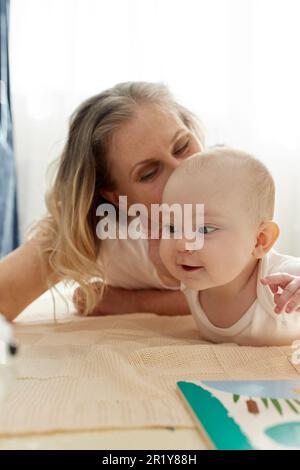 Eine fürsorgliche, fröhliche Mutter spielt mit ihrem kleinen Kind und liegt auf dem Boden in einem hellen Kinderzimmer. Verschiedene Spielzeuge, ein Buch. Spielerisch lernen Stockfoto