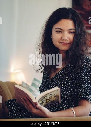 Eine junge asiatische Frau, die Buch liest, während sie auf dem Sofa sitzt Stockfoto