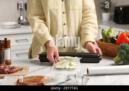 Frau, die eine Schließvorrichtung für Vakuumverpackungen benutzt, mit einer Plastiktüte Zucchini am weißen Tisch in der Küche, Nahaufnahme Stockfoto