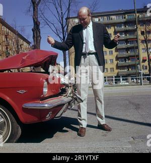 Fahren in der 1960er. Ein Mann steht vor seinem Auto, das bei einem Verkehrsunfall sichtbar beschädigt wurde. Ein Frontalaufprall oder jemand ist in sein Auto zurückgefahren. Er sieht frustriert aus. Schweden 1969 Kristoffersson Stockfoto