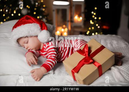 Baby im Weihnachtsschlafanzug und Weihnachtsmannmütze schlafen in der Nähe der Geschenkbox auf dem Bett drinnen Stockfoto