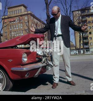 Fahren in der 1960er. Ein Mann steht vor seinem Auto, das bei einem Verkehrsunfall sichtbar beschädigt wurde. Ein Frontalaufprall oder jemand ist in sein Auto zurückgefahren. Er sieht frustriert aus. Schweden 1969 Kristoffersson Stockfoto