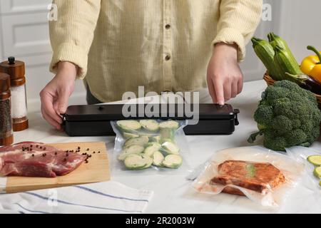 Frau, die eine Schließvorrichtung für Vakuumverpackungen benutzt, mit einer Plastiktüte Zucchini am weißen Tisch in der Küche, Nahaufnahme Stockfoto