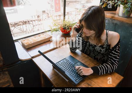 Junge lateinische Transgender-Frau argentinischer Herkunft, die mit ihrem Laptop in einer Cafeteria sitzt, telefoniert, sich auf den Tisch lehnt Stockfoto