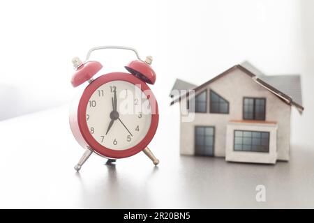 Wecker und Hausmodell. Zeit für eine Hypotheken- oder Immobilienversicherung Stockfoto