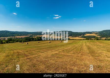 Hügelige Landschaft mit Wiesen, Heuhaufen, unteren meist waldbedeckten Hügeln und kleinen Siedlungen in der tschechischen republik am Sommertag mit blauem Himmel Stockfoto