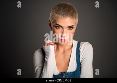 Porträt einer ernsten jungen Frau mit blonden kurzen Haaren, die vor grauem Hintergrund in die Kamera schaut Stockfoto