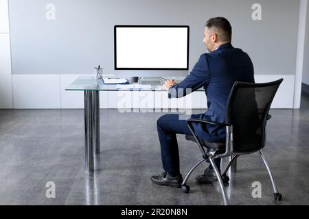 Rückansicht eines Geschäftsmanns im Anzug, der auf einen pc mit leerem Bildschirm für Mock-up schaut, sitzt am Schreibtisch im Büro Stockfoto