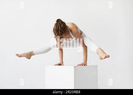 Junge, gesunde Lebensstil Frau, die Yoga praktiziert und eka Pada bhujapidasana Übung, Spinnenhaltung, Handstand auf weißem Würfel, Training in weißen Sportarten Stockfoto