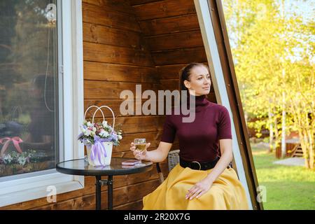 Eine selbstbewusste Frau, die auf der Veranda eines hölzernen Sommerhauses sitzt. Konzept des Sommerurlaubs auf dem Land. Stockfoto